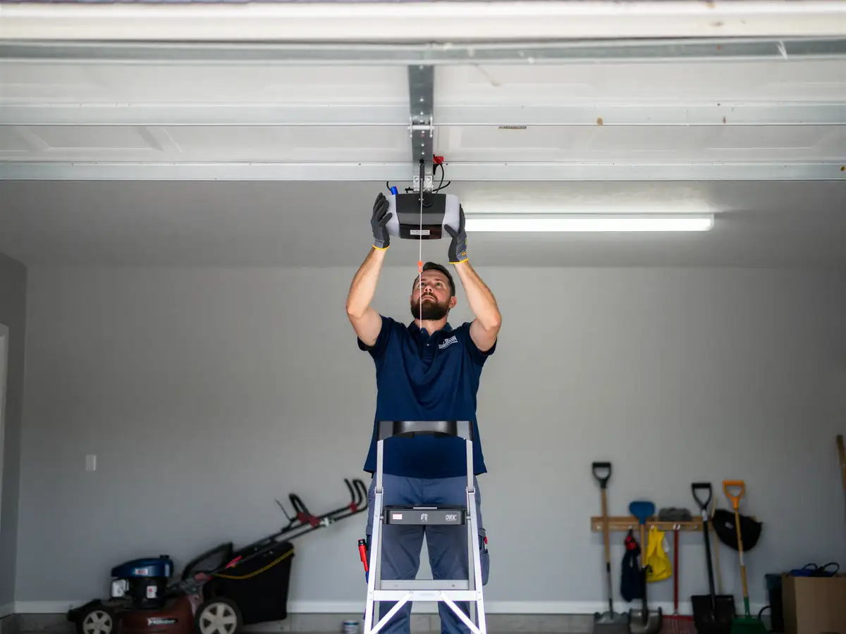 A technician on a ladder installs a garage door opener in a garage.